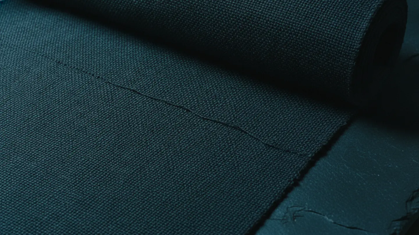 A bolt of machine-woven dark fabric with a single dropped thread, lit by cold cyan raking light on dark slate